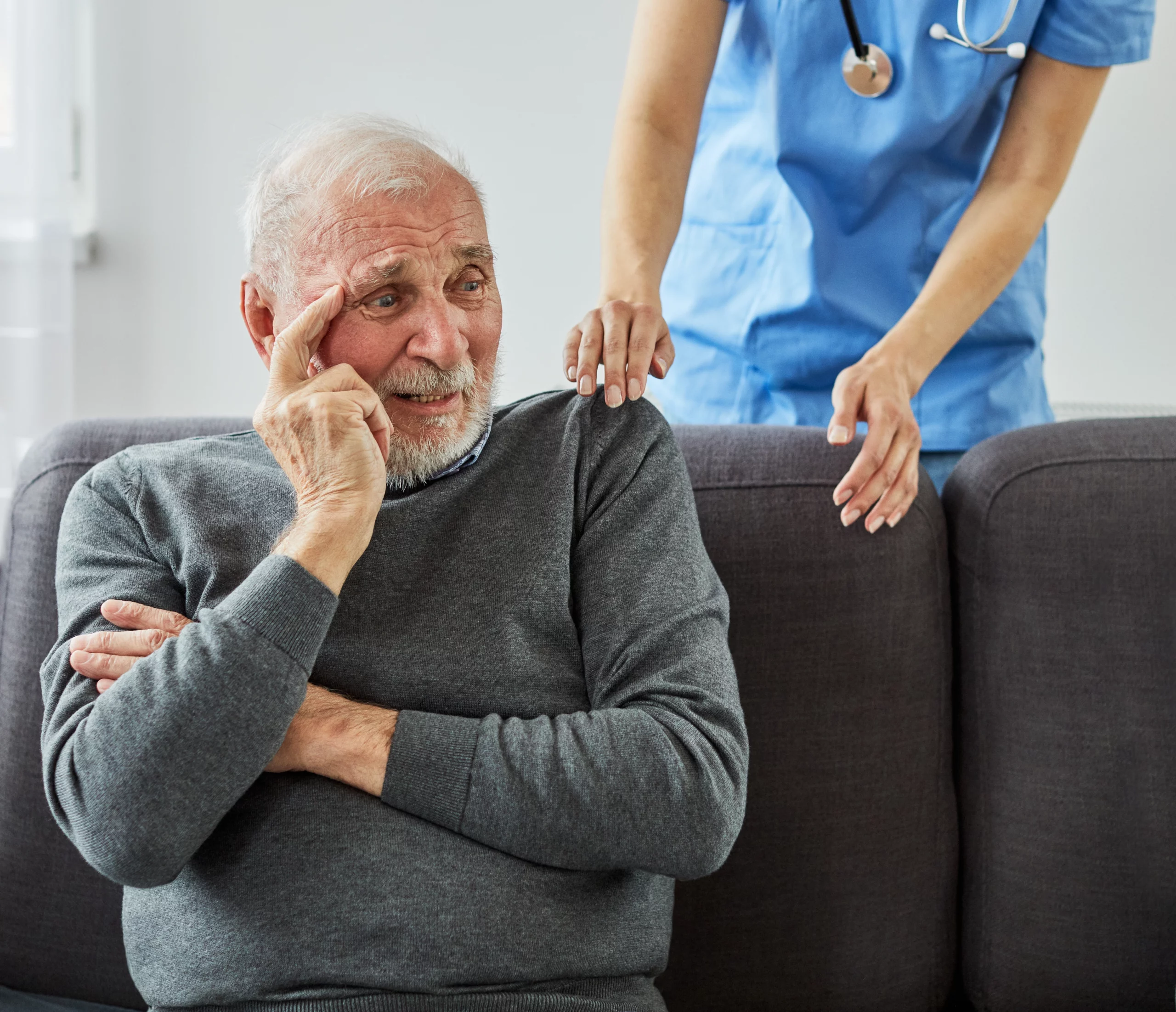 Elderly man in a gray sweater looking pensive on a couch with a nurse in blue scrubs gently supporting his shoulder — conveying care and reassurance in a home health setting.
