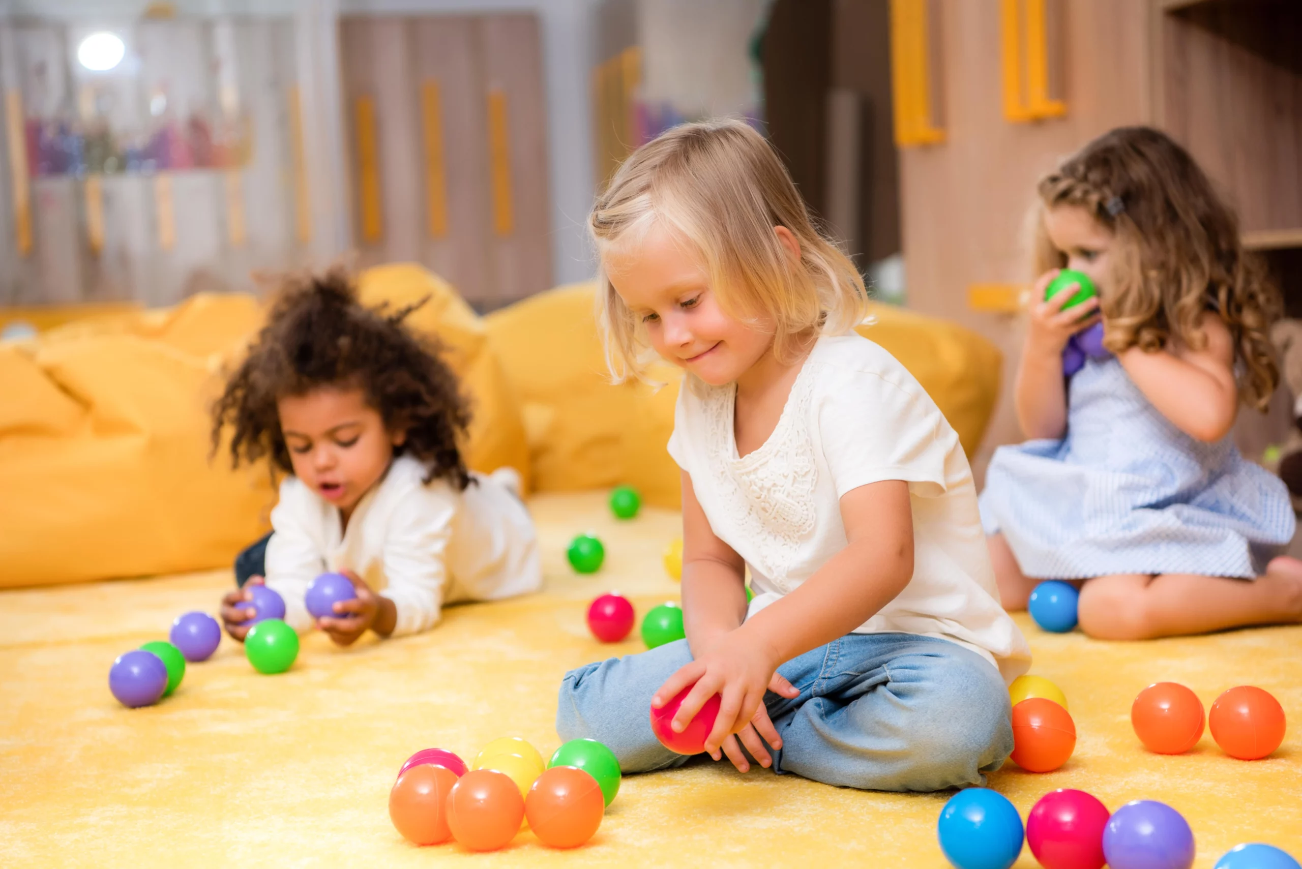 Three young children sitting on a yellow carpet happily playing with colorful plastic balls in a bright, cozy playroom — joyful and carefree.