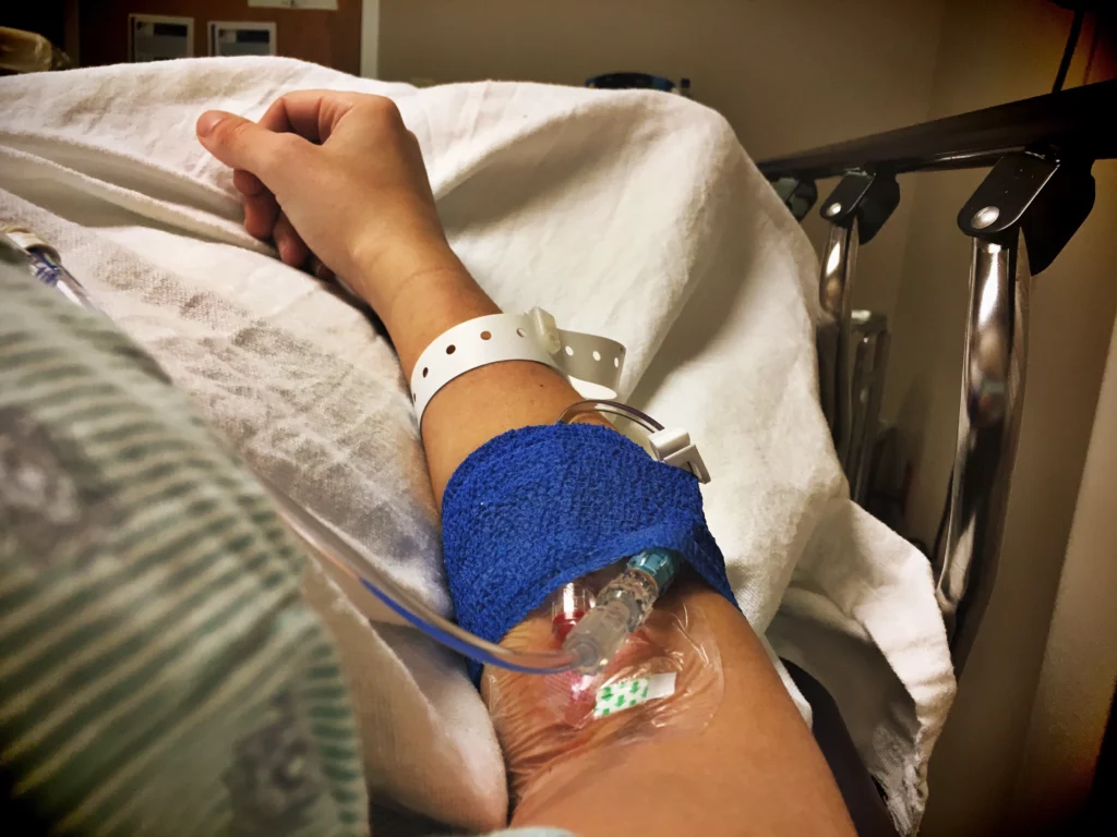 Patient’s arm with an IV line in a hospital bed, wearing a white wristband and resting under a white sheet — conveying a clinical and calm medical environment.
